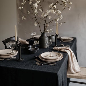 Könnte beinhalten: Eine schwarze Tischdecke mit einem weißen Blumenzweig auf einem beigefarbenen Teller, schwarzen Kerzenständern und einer schwarzen Vase mit weißen Blumen.