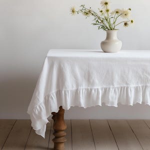 May include: White linen tablecloth with a ruffled edge, draped over a wooden table with a vase of white flowers on top.