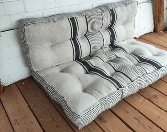 Puede incluir: Un cojín grande, rectangular, a rayas con rayas negras y blancas. El cojín está hecho de una tela de color claro y tiene un diseño acolchado. Está sentado en un suelo de madera.