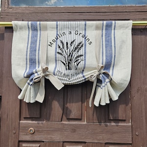 Op de afbeelding: Een witte en blauwe gestreepte stoffen valance met de tekst "Moulin a Grains" en "358 Rue St Denis" in zwart gedrukt. De valance heeft een geplooide vorm en is vastgebonden met beige stoffen banden.