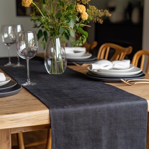 May include: A black linen table runner on a wooden table with a vase of yellow flowers, two wine glasses, and white plates with napkins.