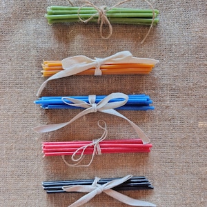 Puede incluir: Conjuntos de velas delgadas y coloridas atadas con cuerda y cintas. Las velas son de color verde, naranja, azul, rojo y negro. Los conjuntos están dispuestos sobre una superficie texturizada de tono neutro.