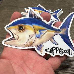 May include: A colorful sticker featuring a stylized fish with blue, yellow, and white details. The fish has an open mouth and the text "SLAPPAFISH" in black. Another sticker is visible in the background.