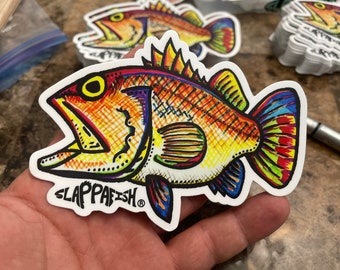 May include: A colorful sticker featuring a stylized fish with an open mouth. The fish is primarily orange with yellow, red, and blue accents. The sticker has the text "SLAPPAFISH" in black. Other similar stickers are visible in the background.