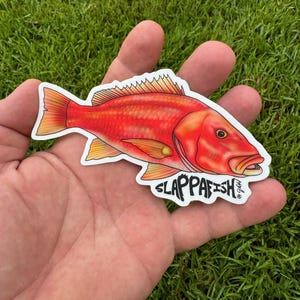 May include: A red fish sticker with the text "SLAPPPAFISH" in black lettering. The sticker is on a hand against a green grass background.