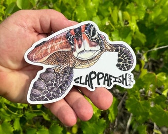 May include: A sticker featuring a detailed illustration of a sea turtle with a brown and green shell, and the text "SLAPPAFISH" in a stylized font. The sticker is held in a hand against a background of green foliage.