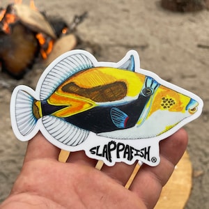 May include: A colorful sticker featuring a detailed illustration of a fish. The fish has yellow, orange, black, and white markings. The sticker has the text "SLAPPAFISH" in white, stylized font.