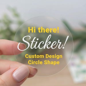 May include: A clear, circular sticker held by a hand. The sticker has the words "Hi there! Sticker!" in white script and yellow block letters. Below, it reads "Custom Design Circle Shape" in yellow.