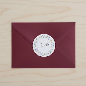 May include: A burgundy envelope with a circular sticker. The sticker has the word "Thanks" in script, surrounded by a wreath of green leaves and small white and purple flowers. The envelope is on a light wood surface.