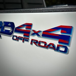 May include: A close-up of a 4x4 Off Road decal with a red, white, and blue American flag design. The decal is on a white surface.