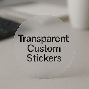 May include: A clear, round sticker with the text "Transparent Custom Stickers" in black. The sticker is slightly frosted and appears to be made of a transparent material. The background is a neutral, light color.