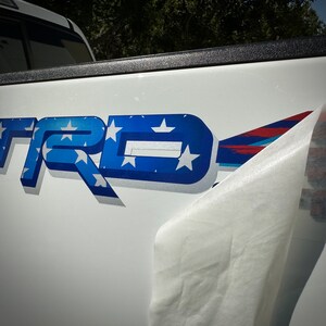 May include: A white truck tailgate with a blue and white "TRD" logo with stars. The logo is a decal that is being peeled off the truck.