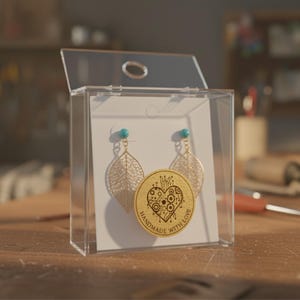 May include: A pair of gold-colored leaf earrings with turquoise studs displayed in a clear acrylic box. A wooden disc with a heart design and the words "HANDMADE WITH LOVE" is also in the box. The earrings are on a white card.