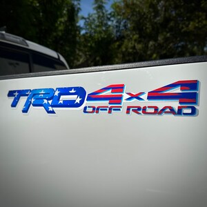 May include: A white truck tailgate with a red, white, and blue "TRD 4x4 OFF ROAD" decal. The decal features a stylized American flag design.