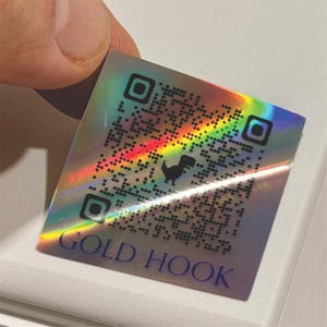 May include: A holographic sticker with a QR code and a dinosaur graphic. The sticker has a rainbow sheen and the words "GOLD HOOK" printed in blue at the bottom. The sticker is held by a person's fingers.