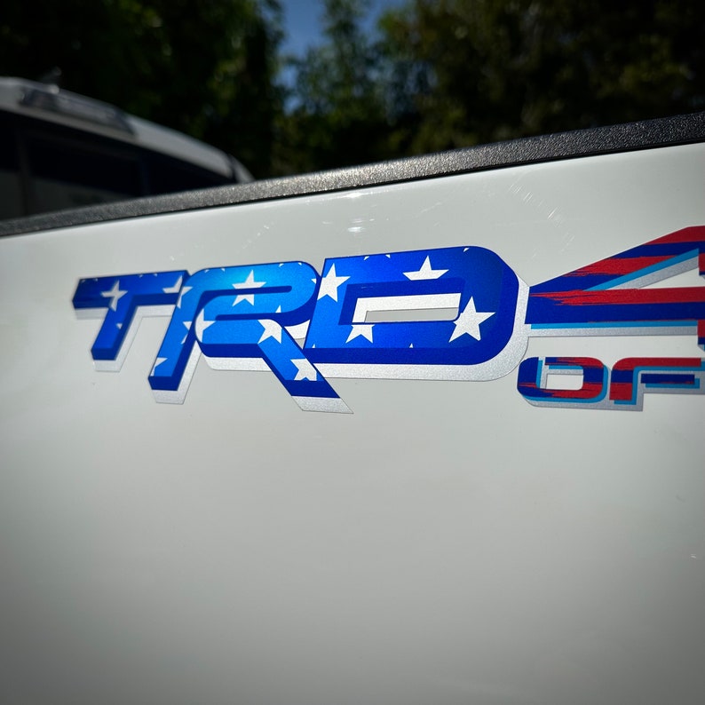 May include: A white truck tailgate with a blue and white "TRD" emblem with stars and a red, white, and blue "4" emblem with the word "OFF" underneath.