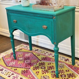 May include: A teal blue painted vintage console table with a single drawer and turned legs. The table is decorated with a vase of pink flowers and a tray with glasses and a pitcher.