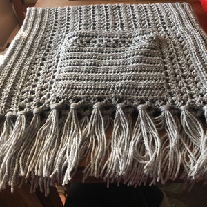 May include: A gray crocheted throw blanket with a fringe border. The blanket has a square design in the center.