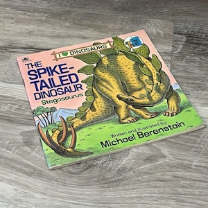 Peut inclure: Un livre pour enfants intitulé The Spike-Tailed Dinosaur Stegosaurus, écrit et illustré par Michael Berenstain. La couverture présente un stégosaure jaune et vert avec de grandes plaques dorsales et une queue épineuse sur un paysage.