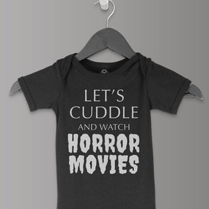 May include: A dark grey baby onesie with short sleeves and a white graphic that says "Let's cuddle and watch horror movies".
