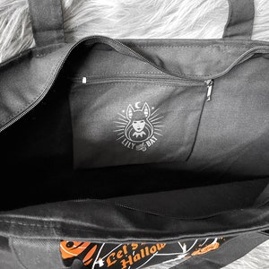 May include: Black canvas tote bag with a zippered pocket featuring a black and white illustration of a woman with bat wings and the text "Lily and Bat". The bag has a black zipper and the text "Let's Hallow" printed on the bottom.