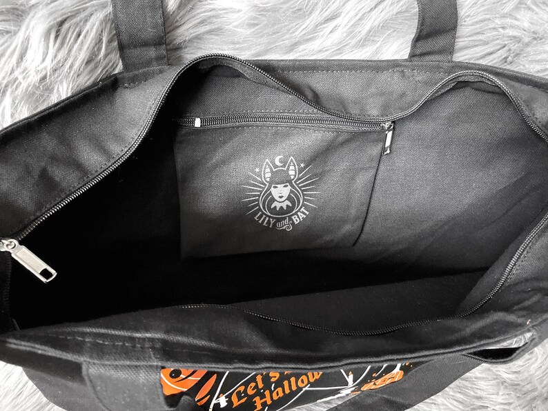 May include: Black canvas tote bag with a zippered pocket featuring a black and white illustration of a woman with bat wings and the text "Lily and Bat". The bag has a black zipper and the text "Let's Hallow" printed on the bottom.