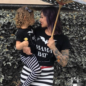 May include: A woman and a child wearing matching black t-shirts with a bat and the text "Mama Bat" and "Baby Bat". The woman is holding a black and white striped umbrella and the child is holding a small yellow flower.