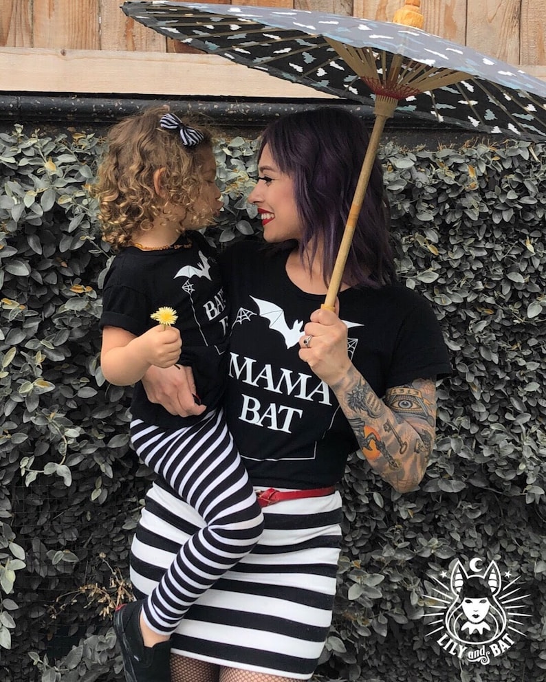 May include: A woman and a child wearing matching black t-shirts with a bat and the text "Mama Bat" and "Baby Bat". The woman is holding a black and white striped umbrella and the child is holding a small yellow flower.