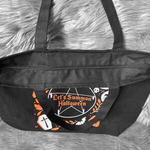 May include: Black canvas tote bag with a zipper closure. The bag has a white and orange design with the text "Let's Summon Halloween" and a pentagram.