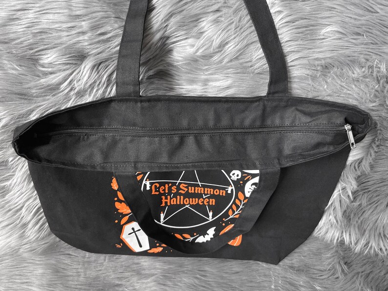 May include: Black canvas tote bag with a zipper closure. The bag has a white and orange design with the text "Let's Summon Halloween" and a pentagram.