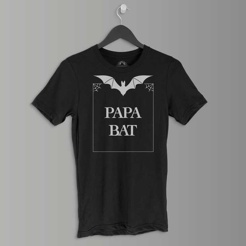Pu&ograve; includere: T-shirt nero con una grafica di pipistrello bianco e il testo "Papa Bat" in un riquadro bianco.