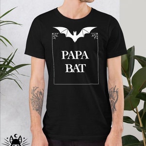 Pu&ograve; includere: Maglietta nera con una grafica di pipistrello bianca e il testo "Papa Bat" in una cornice bianca.