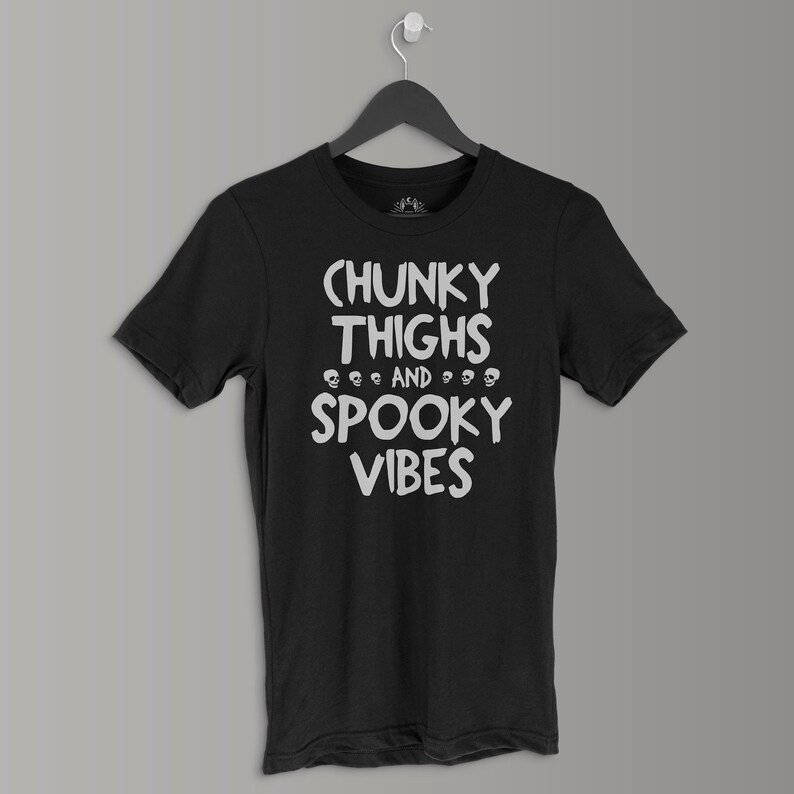 K&ouml;nnte beinhalten: Schwarzes T-Shirt mit wei&szlig;em Text, der "Chunky Thighs and Spooky Vibes" lautet, mit einer Reihe von Totenkopfgrafiken zwischen den W&ouml;rtern.