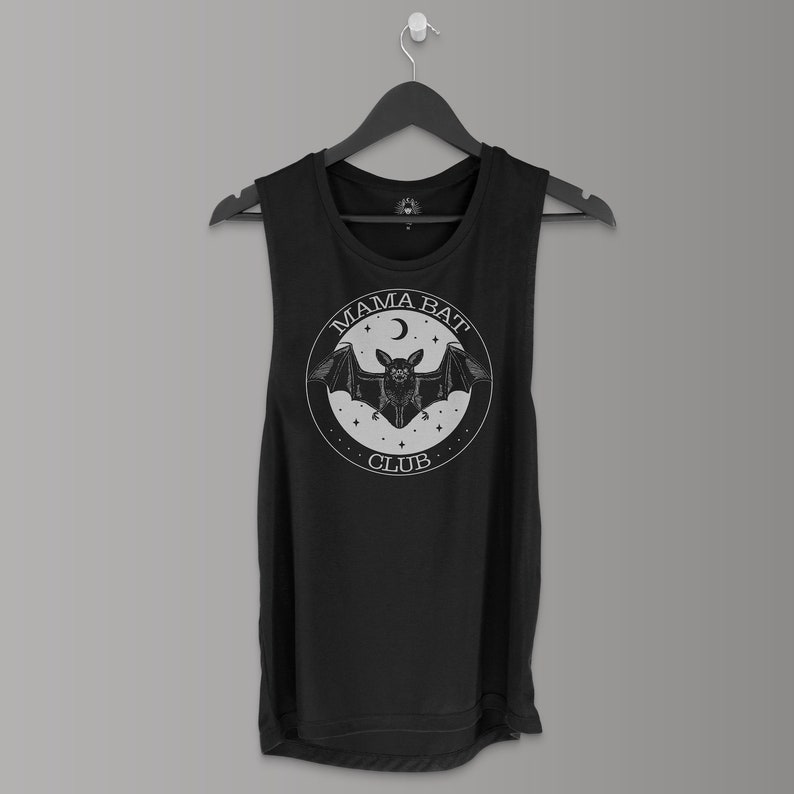 May include: Black sleeveless tank top with a white graphic. The graphic features a bat with outstretched wings inside a circle, with the words "MAMA BAT CLUB" and a crescent moon and stars. The tank top is hanging on a black hanger.
