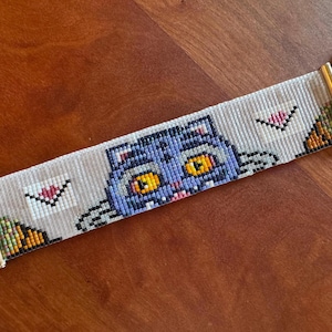 May include: Beaded bracelet featuring a blue cat with yellow eyes and a pink tongue. The cat is surrounded by white envelopes with pink hearts. The bracelet has gold-colored metal ends and a chain.