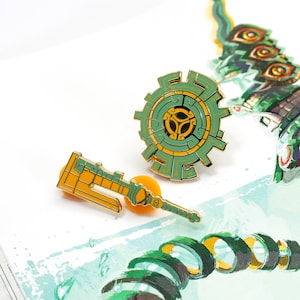 May include: Two gold-colored enamel pins featuring a green and gold shield and a green and gold sword. The shield has a circular design with a central triangle and a maze-like pattern. The sword has a blade with a pointed tip and a handle with a circular design.