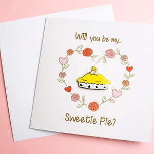Könnte beinhalten: Eine weiße Grußkarte mit dem Text "Will you be my... Sweetie Pie?" zeigt einen herzförmigen Blumenkranz und einen gelben Kuchen mit einem Vogel darauf. Ein weißer Umschlag liegt auf einem rosa Hintergrund.