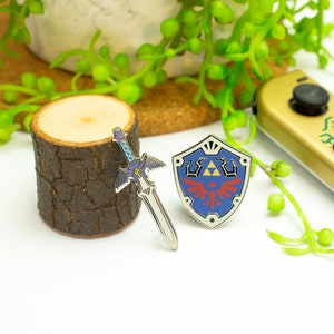May include: A silver and blue enamel pin of the Master Sword from the Legend of Zelda video game series. The sword is on a wooden log with a blue and red enamel pin of the Hylian Shield.