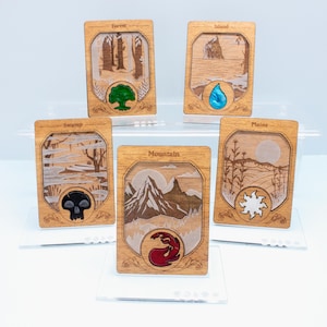 Puede incluir: Cinco tarjetas rectangulares de madera con paisajes grabados a láser e iconos de colores. Las tarjetas están etiquetadas como Forest, Island, Swamp, Mountain y Plains. Cada tarjeta muestra un símbolo único: un árbol verde, una gota de agua azul, una calavera negra, una llama roja y un sol blanco.
