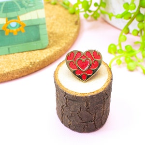 Könnte beinhalten: Goldene herzförmige Anstecknadel mit rotem Emaille-Design. Die Anstecknadel hat ein kleineres Herz im Inneren des größeren Herzens. Die Anstecknadel liegt auf einem kleinen Holzscheit.
