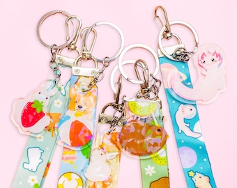 May include: Five colorful keychains with different charms and designs. Each keychain has a silver metal ring and a colorful fabric strap. The charms include a strawberry, a corgi, a kiwi, a pineapple, and a cartoon animal.