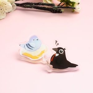 May include: Two clear acrylic pins with cartoon bird designs. One shows a light blue pigeon on a yellow cloud. The other depicts a black bird with a red accent and question marks. The pins are set against a pink backdrop.