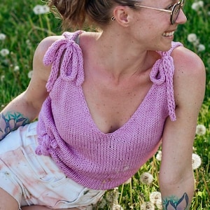 Könnte beinhalten: Eine Frau trägt ein helllilafarbenes Stricktop mit einem Bindedetail vorne. Sie sitzt in einem Feld mit grünem Gras und weißen Blumen. Sie hat ein rosa Haargummi im Haar.
