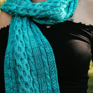 May include: A teal blue knitted scarf with a cable knit pattern. The scarf is draped around the shoulders of a person wearing a black off-the-shoulder top.