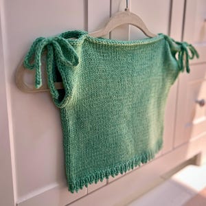 Könnte beinhalten: Ein hellgrünes Strickoberteil mit Trägern zum Binden hängt an einem beigen Kleiderbügel. Das Oberteil hat einen gewellten Saum und wird vor einem weißen Schrank mit floralen Knöpfen präsentiert. Der Strickstoff hat ein strukturiertes Aussehen.