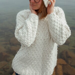 Puede incluir: Una mujer con un su&eacute;ter de punto blanco y un gorro de punto blanco con un pomp&oacute;n de piel marr&oacute;n. Lleva gafas redondas y tiene las manos en las mejillas.