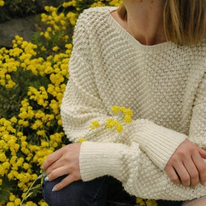 May include: A close-up of a white knitted sweater with a textured pattern. The sweater is worn by a person sitting in a field of yellow flowers.