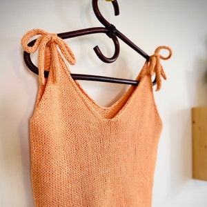 May include: A peach-colored knitted tank top with a V-neck and tie straps. The tank top is hanging on a wooden hanger.