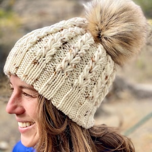 Könnte beinhalten: Eine weiße Strickmütze mit einem braunen Kunstfellbommel. Die Mütze hat ein Zopfmuster.