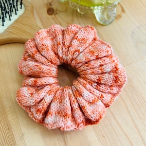 Può includere: Un elastico per capelli lavorato a maglia in una combinazione di colori pesca e bianco. L'elastico è realizzato con un filato morbido e testurizzato e ha un bordo arricciato.